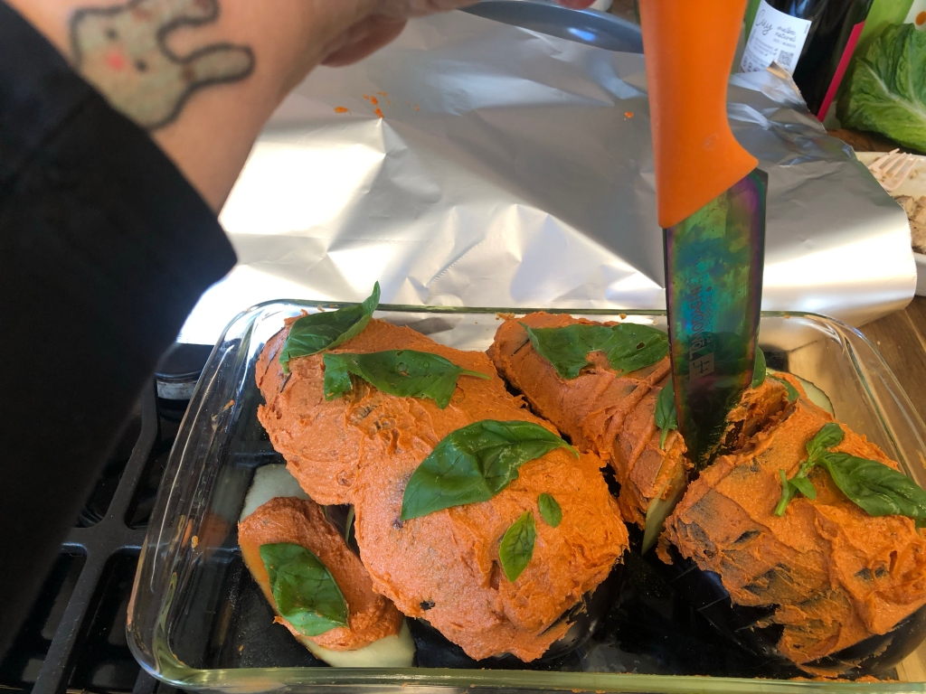 Sitting in a glass 9x12 pan in front of a piece of aluminum foil are two eggplants covered in a red sauce and whole basil leaves with a knife sticking between two of the slices to show that you should push the sauce down in.